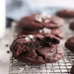Freshly baked chocolate cookies with sea salt on a cooling rack next to a glass of milk.