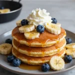 A stack of golden banana pancakes topped with fresh banana slices and syrup on a white plate.