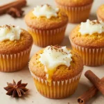 Freshly baked pumpkin cream cheese muffins on a wooden table with a bowl of cream cheese frosting nearby.