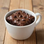 A rich chocolate brownie served in a white mug, topped with a scoop of vanilla ice cream.