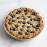 A close-up of a baked blueberry muffin oatmeal in a white dish, topped with fresh blueberries and a drizzle of honey.