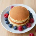 A stack of golden protein pancakes served on a plate, topped with fresh fruit and syrup.