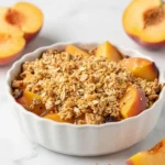 A close-up of a freshly baked peach crisp with a golden-brown cinnamon-oat topping in a white baking dish.
