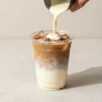 A glass of iced pumpkin chai latte with ice cubes, whipped cream, and cinnamon sprinkled on top, served on a kitchen table