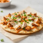 Buffalo chicken flatbread topped with shredded chicken, melted cheese, and drizzled buffalo sauce on a baking tray.