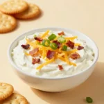 A creamy million dollar crack dip topped with shredded cheese, bacon crumbles, and chopped green onions served in a white bowl with crackers on the side