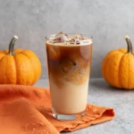 A glass of pumpkin cold brew coffee topped with creamy foam and a sprinkle of pumpkin spice on a wooden table.