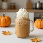 A pumpkin spice latte served iced in a clear glass with whipped cream, cinnamon, and pumpkin spice sprinkle on a rustic table