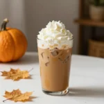 A frothy iced pumpkin spice latte in a clear glass with ice cubes, cinnamon, and whipped cream beside a warm hot pumpkin spice latte in a mug.