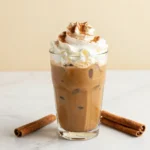 A glass of pumpkin spice cold brew topped with creamy foam and a sprinkle of cinnamon on a wooden table.